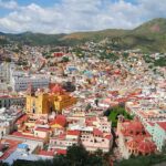 An expansive panoramic view of Guanajuato City, Mexico, showcasing its vibrant, colorful houses covering rolling hills, with prominent landmarks like the yellow Basílica Colegiata de Nuestra Señora de Guanajuato and other historic buildings, under a partly cloudy sky.