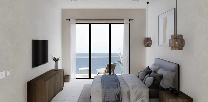 A modern bedroom with white walls features a bed with grey and blue bedding, a wall-mounted TV, and a sliding glass door leading to a balcony with an ocean view.